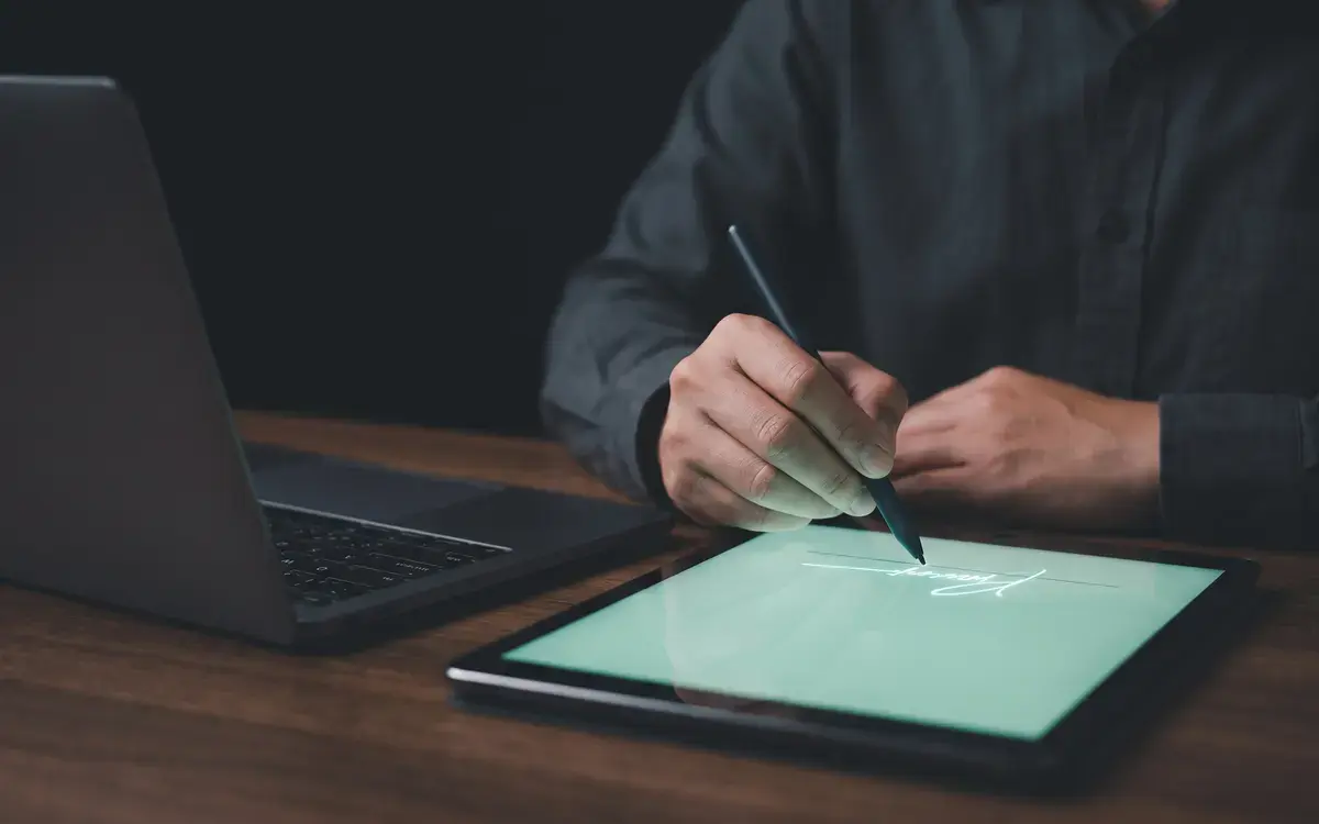Une personne signe un document sur une tablette avec un stylet, un ordinateur portable à côté.