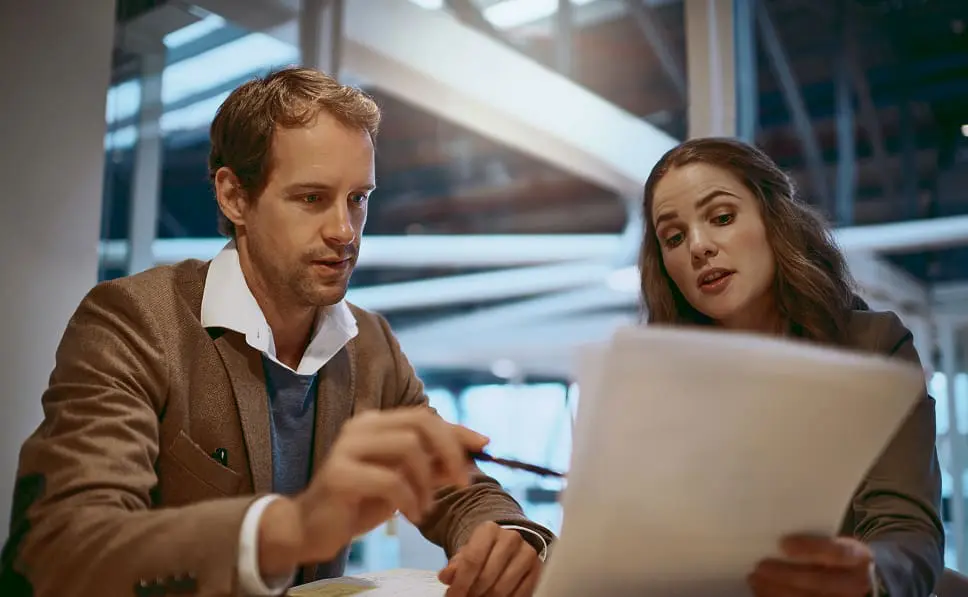 A man and a woman sit at a desk in an office, reviewing documents related to managed cloud services. The man is holding a pen and pointing at the paper, while the woman looks at the document and explains something.