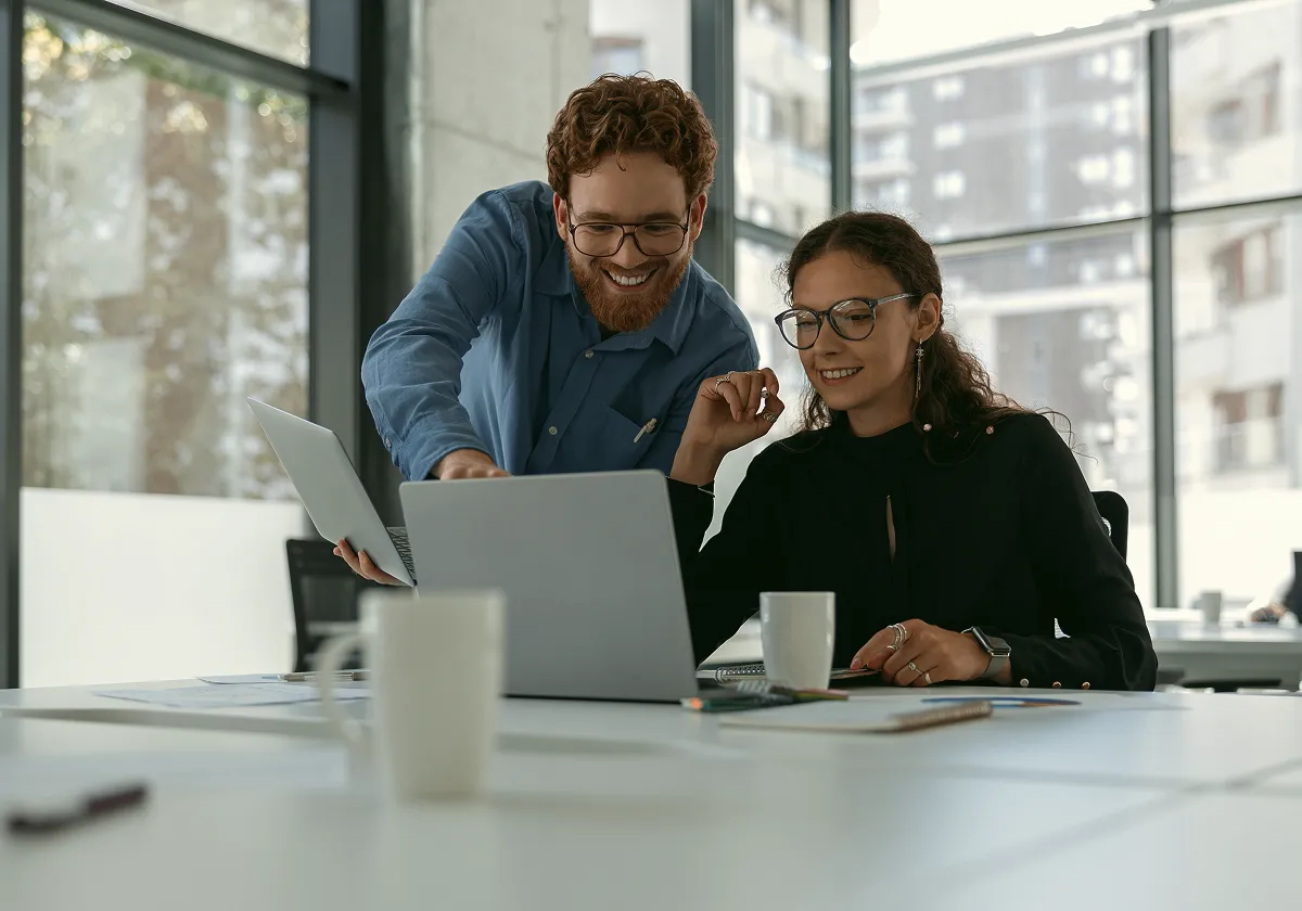 Two colleagues in an office, one standing and pointing at a laptop screen while the other sits and smiles. Both wear glasses and appear to be discussing work, with notebooks and coffee mugs on the desk.