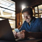 A smiling man sits at a desk in a modern office, looking at his mobile whilst his laptop is open in front of him. He appears happy and engaged, with warm lighting illuminating the workspace.