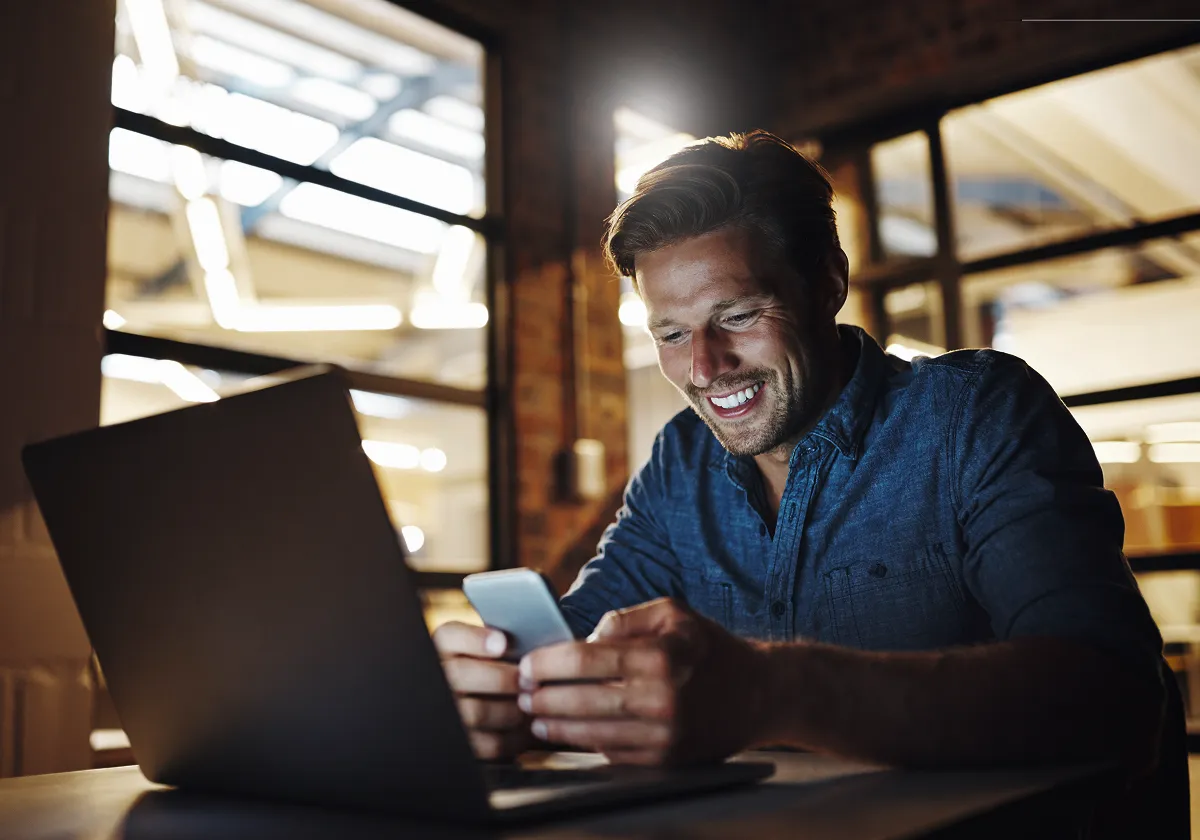 A smiling man sits at a desk in a modern office, looking at his mobile whilst his laptop is open in front of him. He appears happy and engaged, with warm lighting illuminating the workspace.
