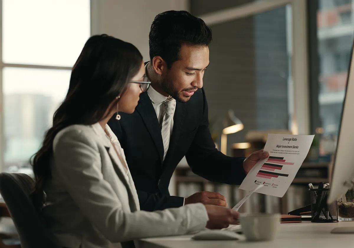 Two business professionals in an office review a document with colourful bar graphs. The man points to the chart whilst the woman listens attentively, both dressed in business attire and seated at a desk with a computer.