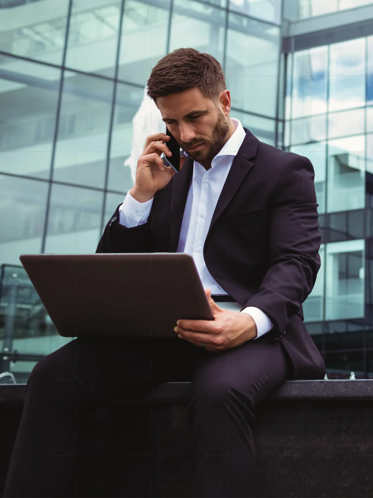 A man in a black suit sits outdoors in front of a modern glass building, talking on his mobile while working on a laptop, appearing focused and professional.