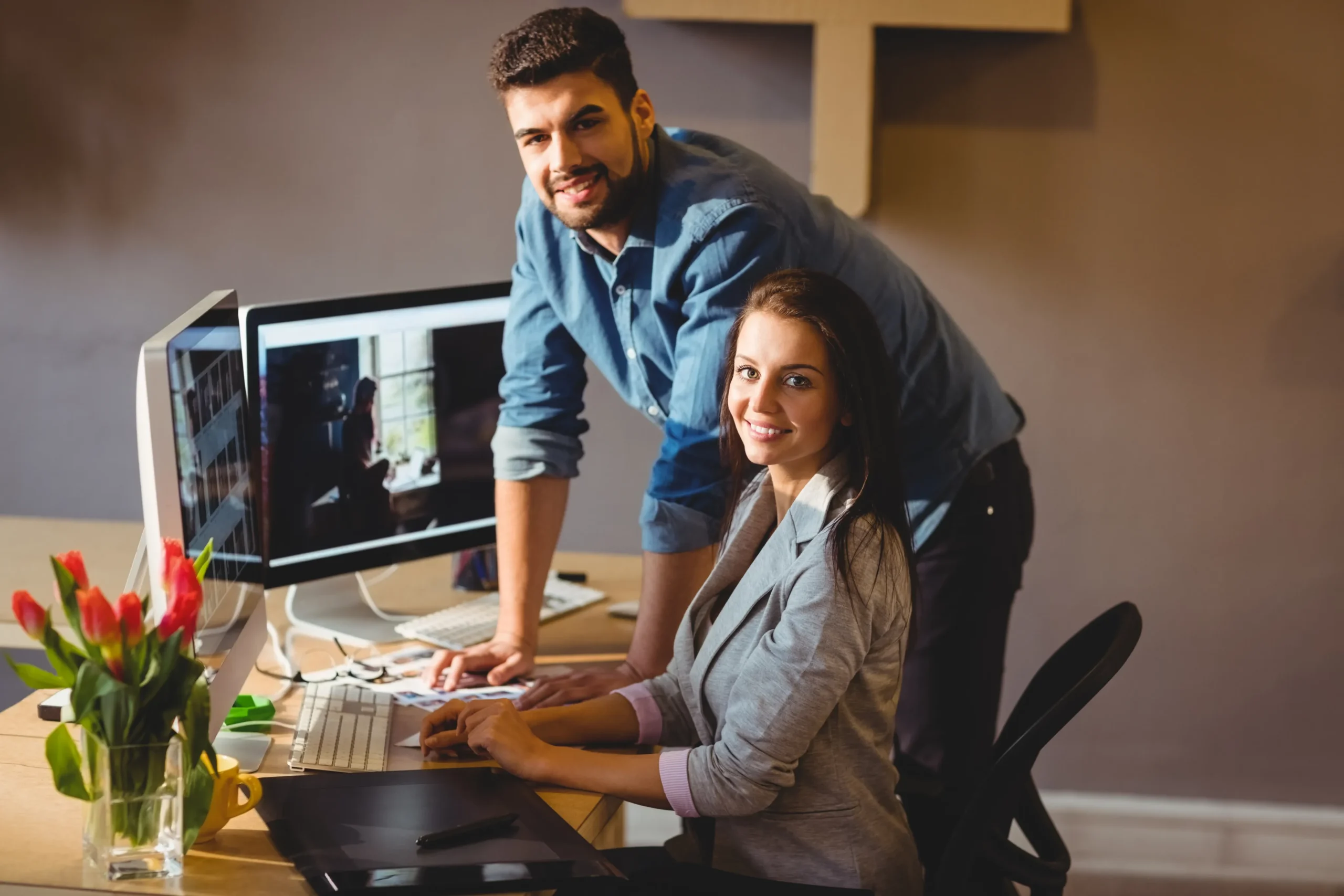 A man and a woman smile at the camera while working at a computer desk. The woman is seated and typing, whilst the man stands beside her. There are two monitors and a vase of red tulips on the desk.