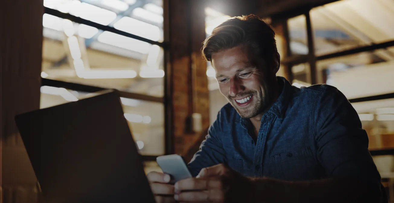 A man in a denim shirt sits at a desk with a laptop, smiling whilst looking at his mobile phone in a warmly lit modern office or workspace.